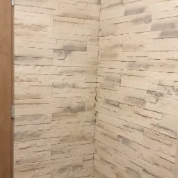 Fragment ściany wykończonej dekoracyjnym kamieniem w odcieniach beżu i szarości, widoczny narożnik i fragment drewnianej ościeżnicy drzwiowej po lewej stronie.