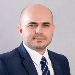 Portret mężczyzny w granatowym garniturze i krawacie w paski, z lekkim uśmiechem na tle szarej ściany.