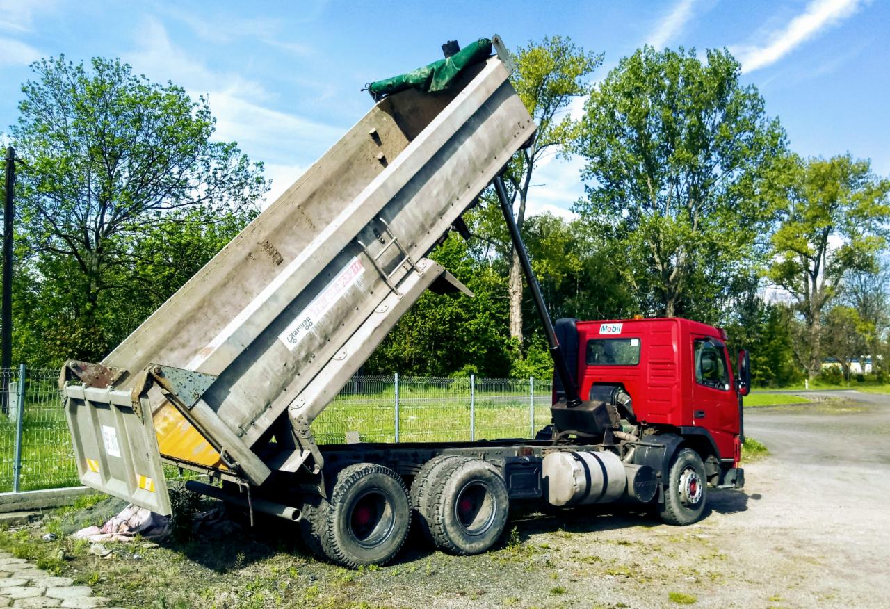 Czerwona ciężarówka wywrotka z podniesioną skrzynią ładunkową na tle zieleni drzew i błękitnego nieba.