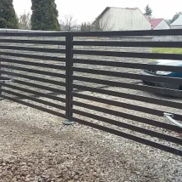 Automatyczna brama wjazdowa z poziomymi profilami, zamontowana na kamiennych słupkach, z widocznym mechanizmem otwierającym i podjazdem wysypanym żwirem.