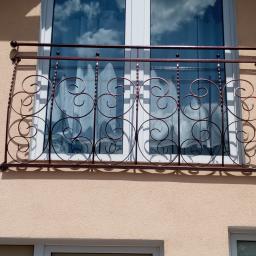 PORTAM Zwoliński - Ozdobna balustrada balkonowa z metalu w kolorze miedzianym, z motywem spiralnym, zamontowana na balkonie z oknem z białymi ramami, w tle odbicie nieba w szybach.