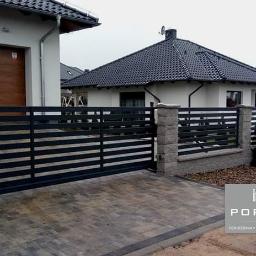 PORTAM Zwoliński - Nowoczesne, poziome przęsła metalowe tworzą ogrodzenie posesji z bramą przesuwną, uzupełnione kamiennymi słupkami i kostką brukową na podjeździe.