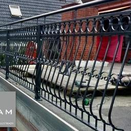 PORTAM Zwoliński - Czarna, metalowa balustrada balkonowa z dekoracyjnymi elementami w kształcie kółek, zamontowana na balkonie z ceglaną ścianą w tle.