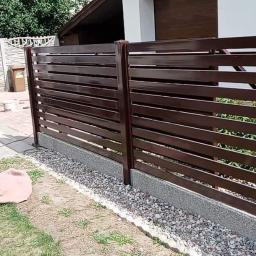 PORTAM Zwoliński - Nowoczesne, poziome, brązowe przęsła ogrodzeniowe z metalu, zamontowane na betonowej podmurówce z kamiennym podsypem, widoczne na posesji w Różankach. W tle brama wjazdowa i zaparkowany bus.