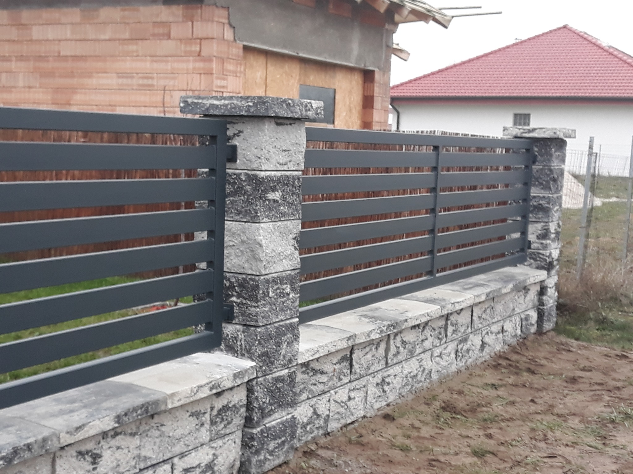 Nowoczesne ogrodzenie z poziomych, szarych profili metalowych, osadzone na niskim murku z szarego kamienia łupanego, z słupkami z tego samego kamienia, w tle budynek w trakcie budowy.