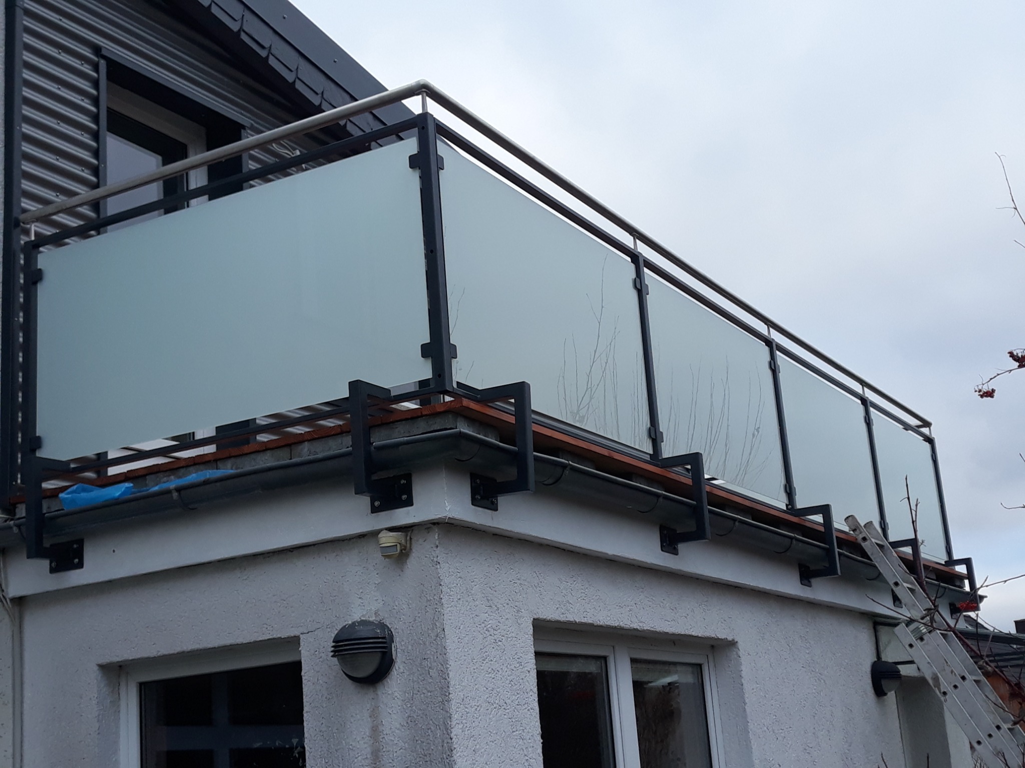Barierka balkonowa z matowego szkła w czarnej metalowej ramie i z elementami ze stali nierdzewnej, widok z dołu, drabina oparta o ścianę budynku.