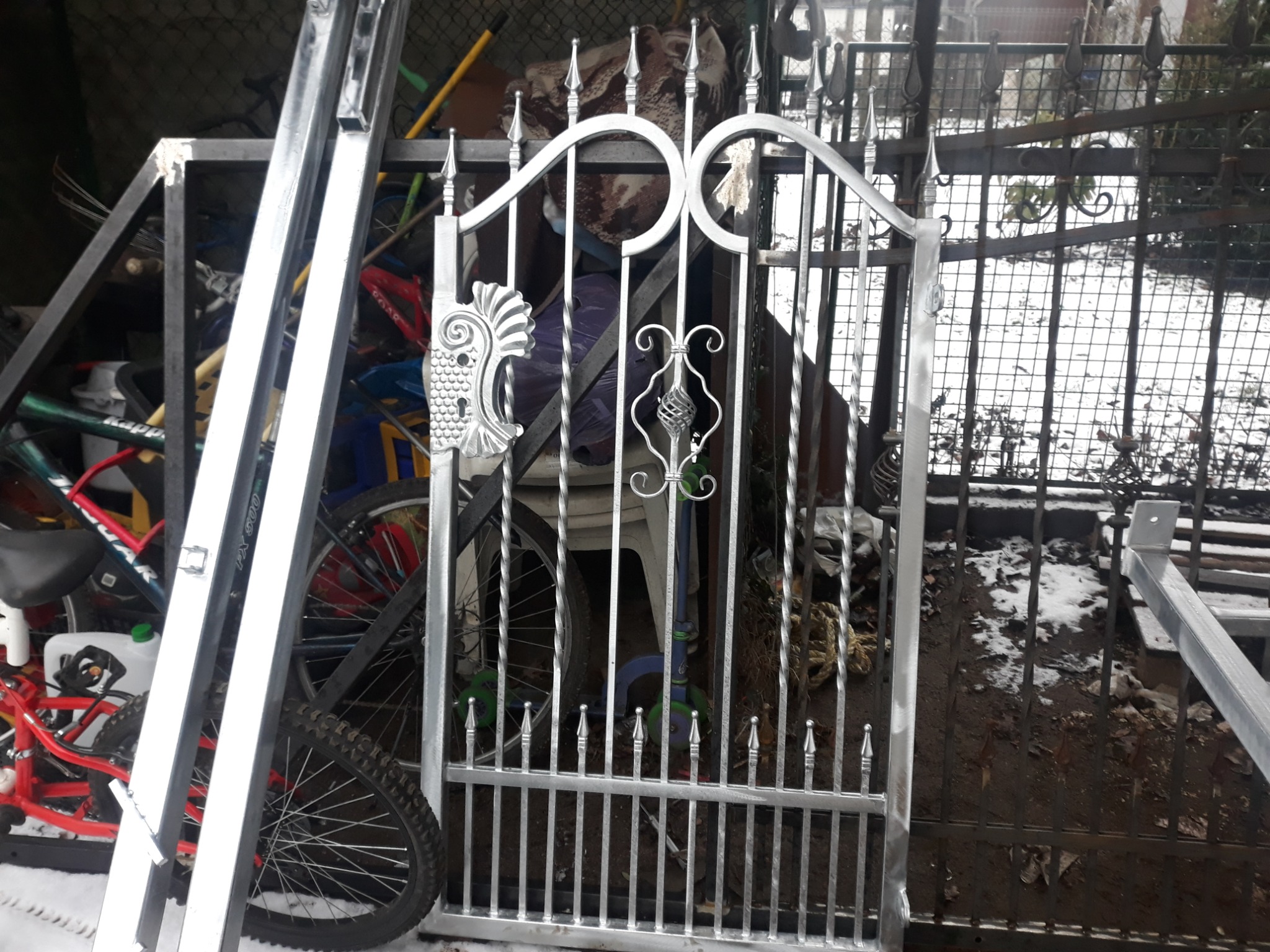 Nieskończona, srebrna, dekoracyjna brama z kutego żelaza, oparta o rower i drabinę, w tle widoczny ogród pokryty śniegiem.