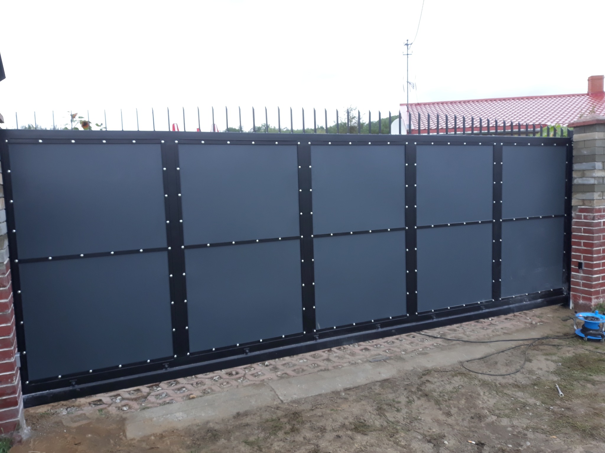 Nowoczesna, przesuwna brama wjazdowa z szarymi panelami i czarną ramą, zabezpieczona kolcami na górze, widoczna z zewnątrz posesji, z boku mur z czerwonej cegły i zwinięty przedłużacz.