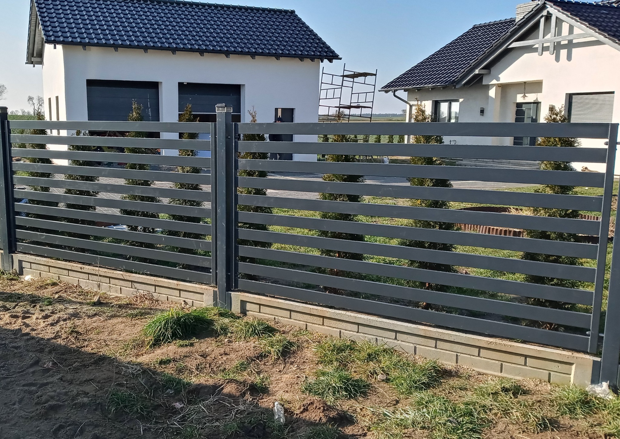 Nowoczesne ogrodzenie panelowe w kolorze grafitowym z poziomymi profilami, zamontowane na niskiej podmurówce z cegły, w tle dwa domy jednorodzinne i młode tuje.