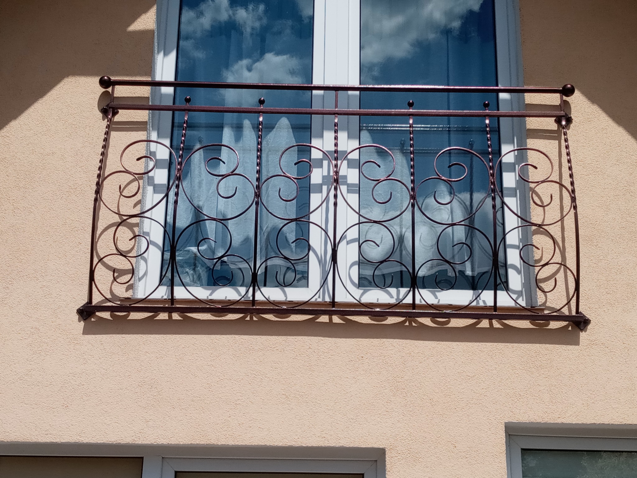 Ozdobna balustrada balkonowa z metalu w kolorze miedzianym, z motywem spiralnym, zamontowana na balkonie z oknem z białymi ramami, w tle odbicie nieba w szybach.