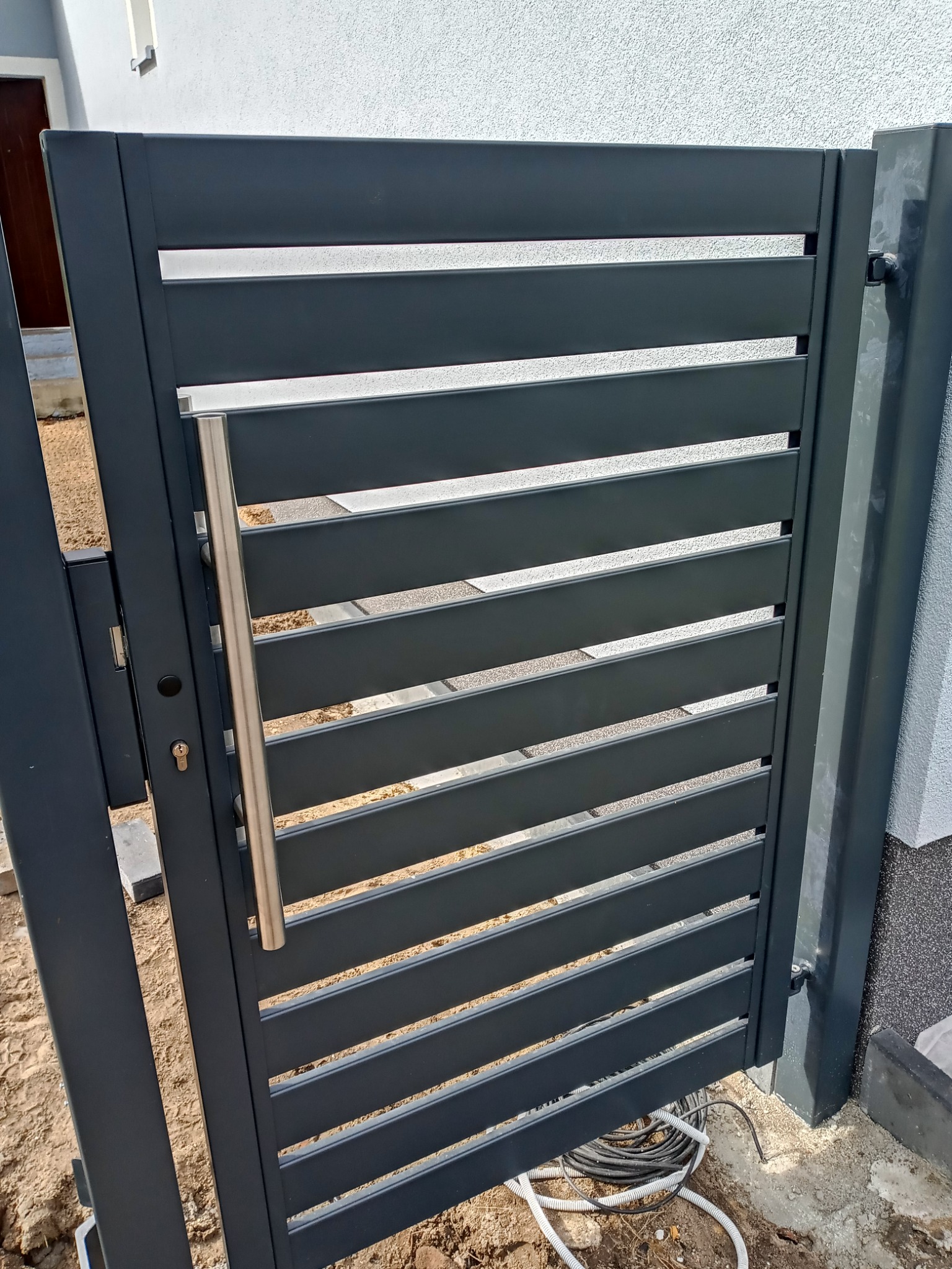 Nowoczesna, pozioma brama w kolorze grafitowym z metalowym uchwytem, widoczny zamek i przewody elektryczne na ziemi.