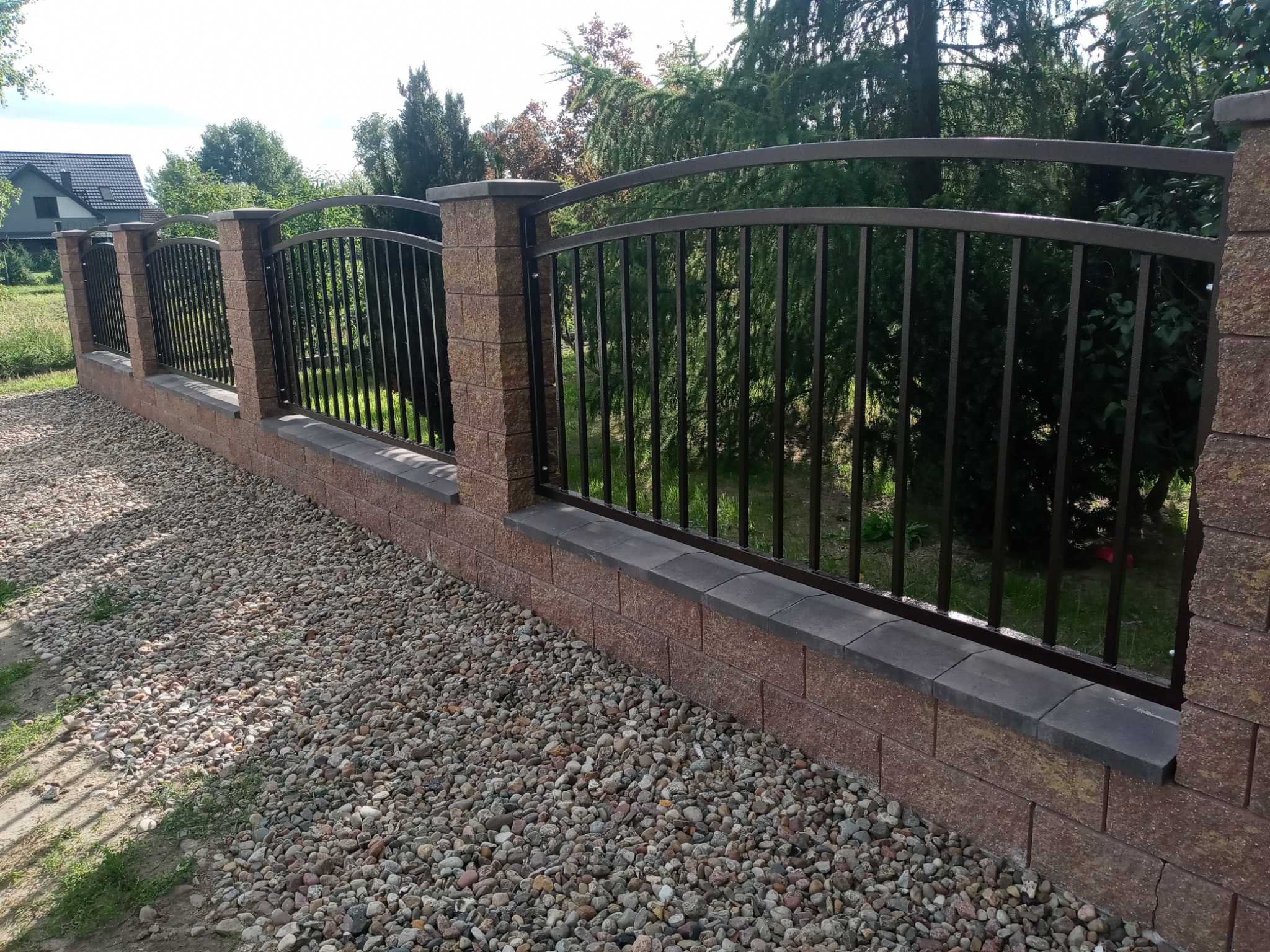 Nowoczesne ogrodzenie z metalowymi prętami i ceglanymi słupkami, wykończone szarymi elementami, otoczone żwirem i zielenią.
