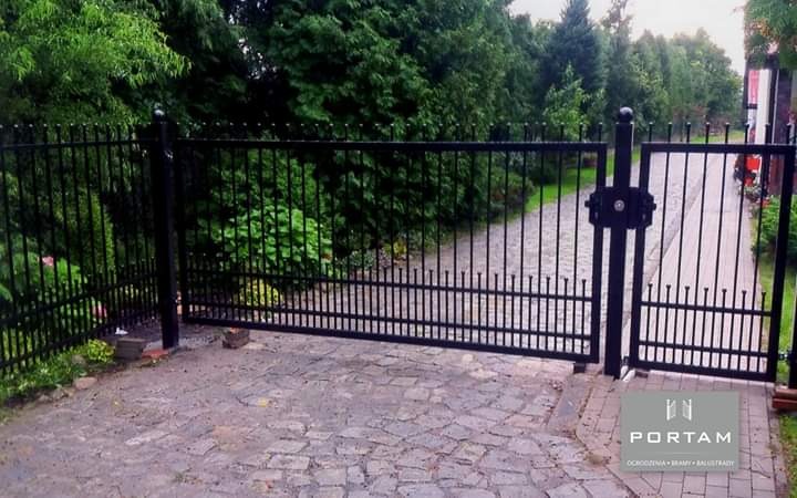 Otwarta, czarna, metalowa brama wjazdowa z kutymi elementami na posesję z brukowanym podjazdem, widoczny logo firmy Portam.