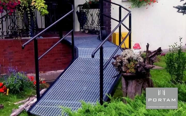 Metalowy podjazd dla wózków inwalidzkich z czarną balustradą, prowadzący do wejścia domu, z logo firmy Portam w prawym dolnym rogu, otoczony zielenią.