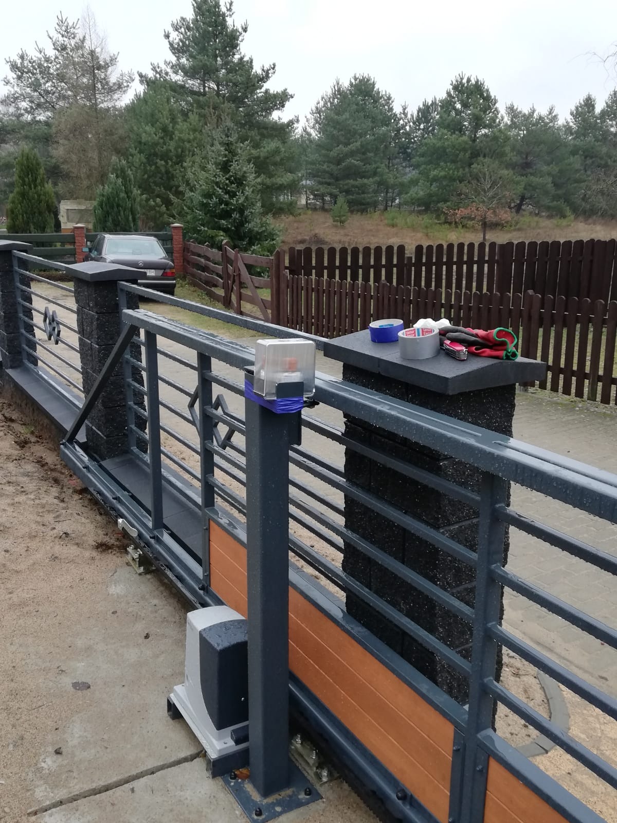 Szara, przesuwna brama wjazdowa z automatyką, widoczne narzędzia i taśmy na słupku, tło z drzewami i samochodem.