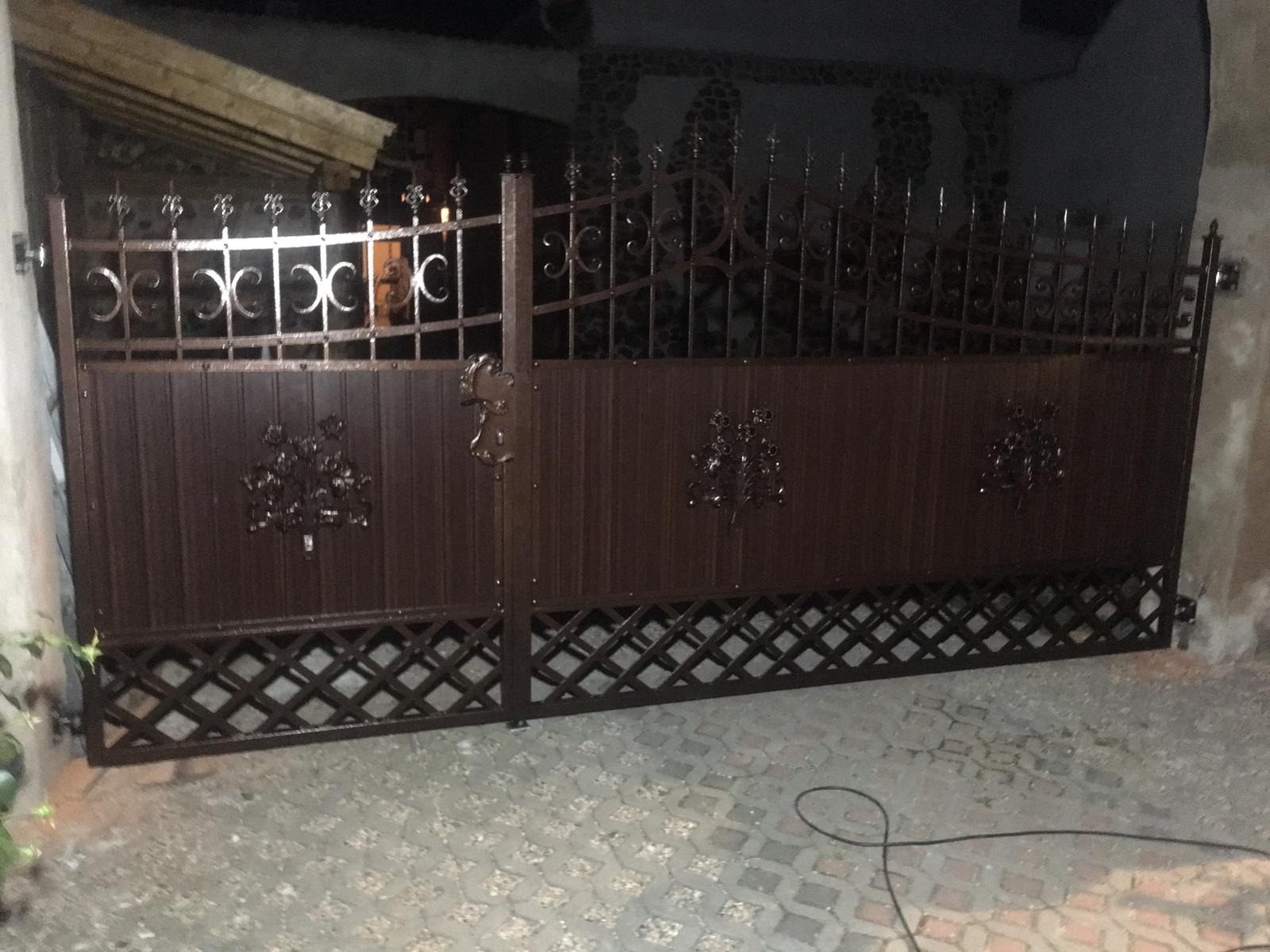 Brunatna brama wjazdowa z dekoracyjnymi aplikacjami w kształcie drzewek i górną częścią z elementami ozdobnymi w kształcie półksiężyców, widoczna na tle ściany z mozaiką kamienną.