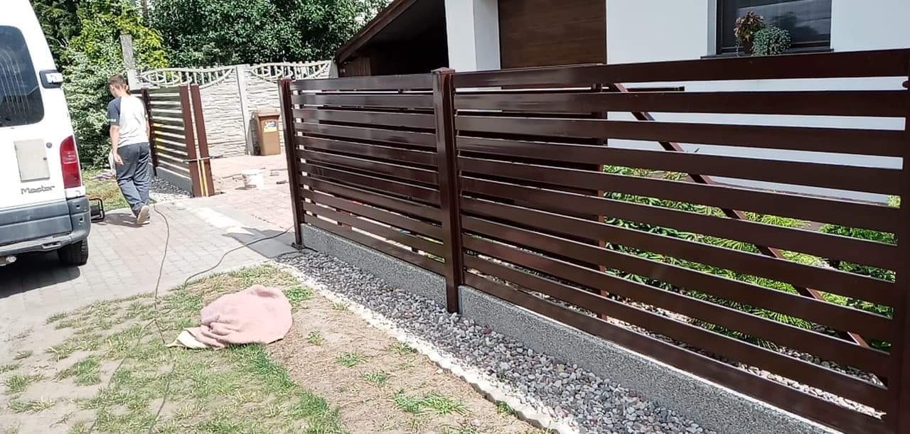 Nowoczesne, poziome, brązowe przęsła ogrodzeniowe z metalu, zamontowane na betonowej podmurówce z kamiennym podsypem, widoczne na posesji w Różankach. W tle brama wjazdowa i zaparkowany bus.