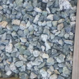 Krawędź drewnianej skrzynki wypełniona drobnymi, szaro-zielonymi kamieniami o nieregularnych kształtach i białych przebarwieniach.