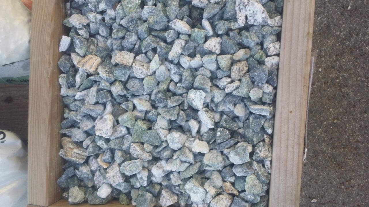 Krawędź drewnianej skrzynki wypełniona drobnymi, szaro-zielonymi kamieniami o nieregularnych kształtach i białych przebarwieniach.