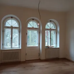 Pomalowane na biało wnętrze z drewnianą podłogą, trzema oknami łukowymi i grzejnikami. Na parapecie okiennym widoczne wałki do malowania i puszka farby.