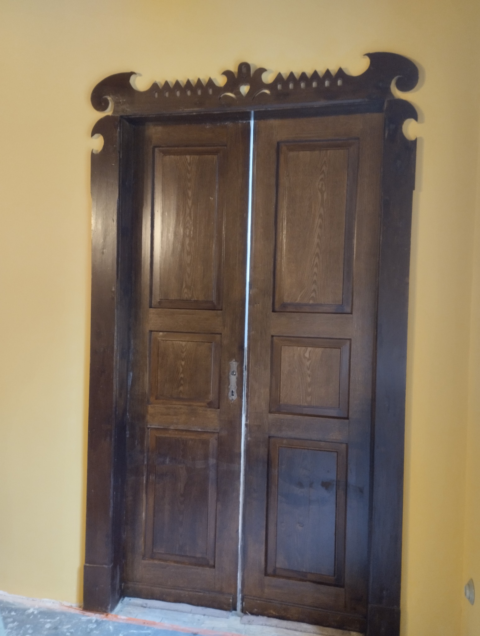 Drewniane, dwuskrzydłowe drzwi z ozdobną ramą w stylu retro, po renowacji, na tle żółtej ściany.