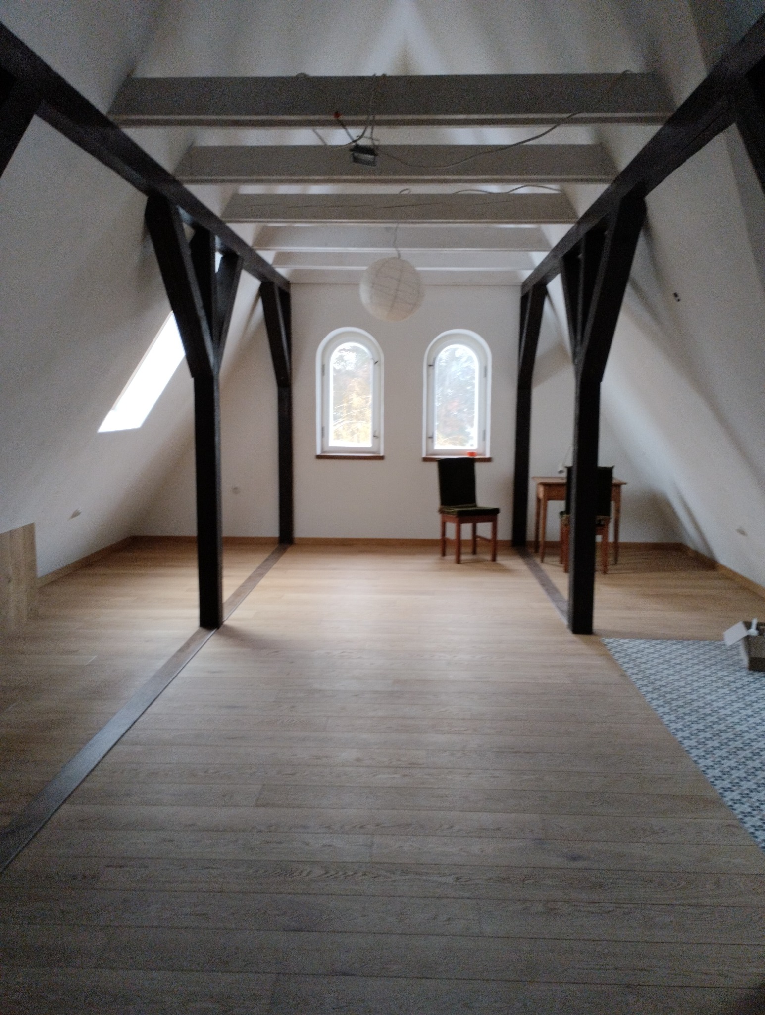 Poddasze po remoncie z drewnianą podłogą, widoczne odsłonięte drewniane belki konstrukcyjne, dwa okna w kształcie łuku i fragment dywanu w rogu pomieszczenia.