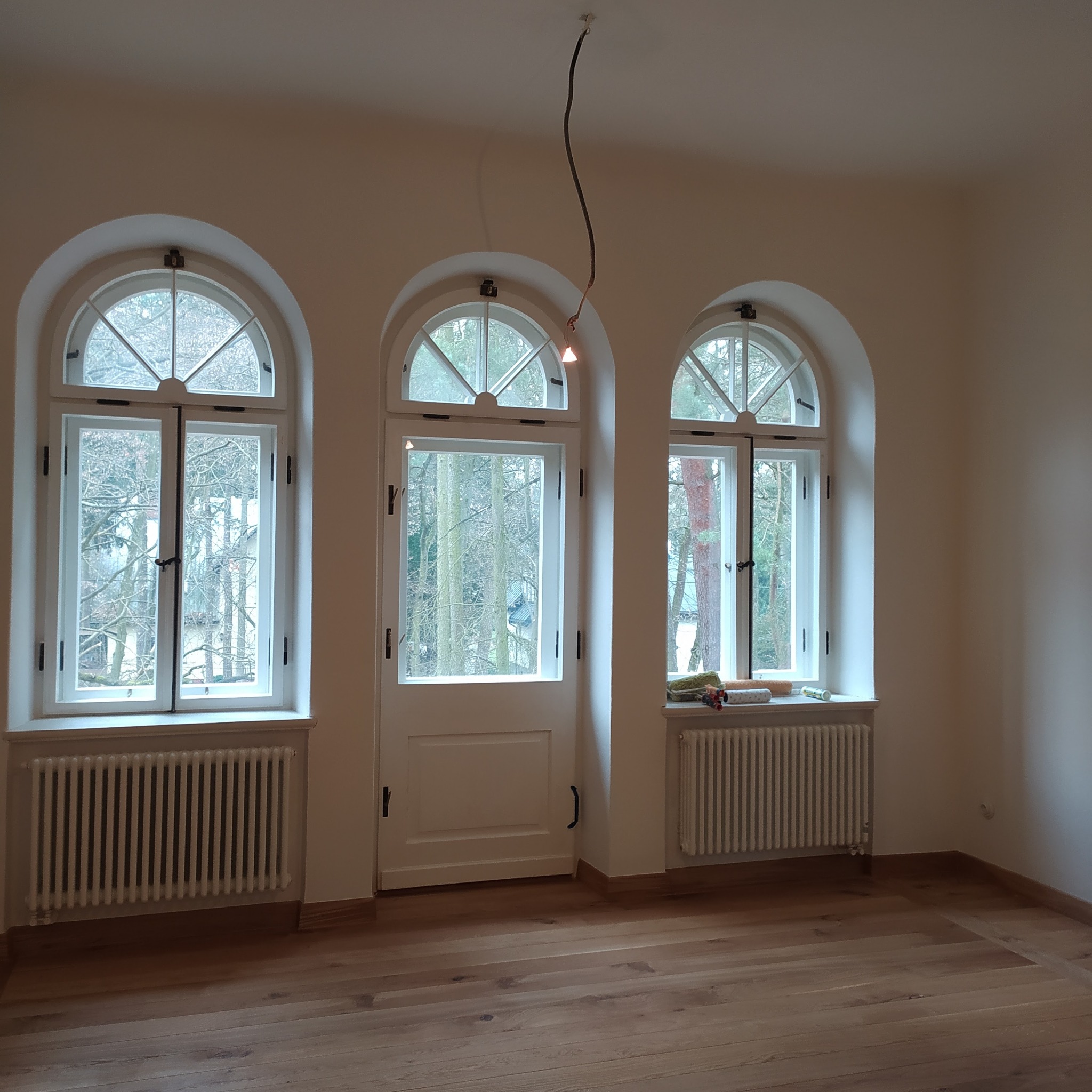 Pomalowane na biało wnętrze z drewnianą podłogą, trzema oknami łukowymi i grzejnikami. Na parapecie okiennym widoczne wałki do malowania i puszka farby.
