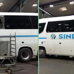 Pracownik na rusztowaniu montuje szybę w autokarze Sindbad, zabezpieczonym taśmą, widok z boku na warsztat.