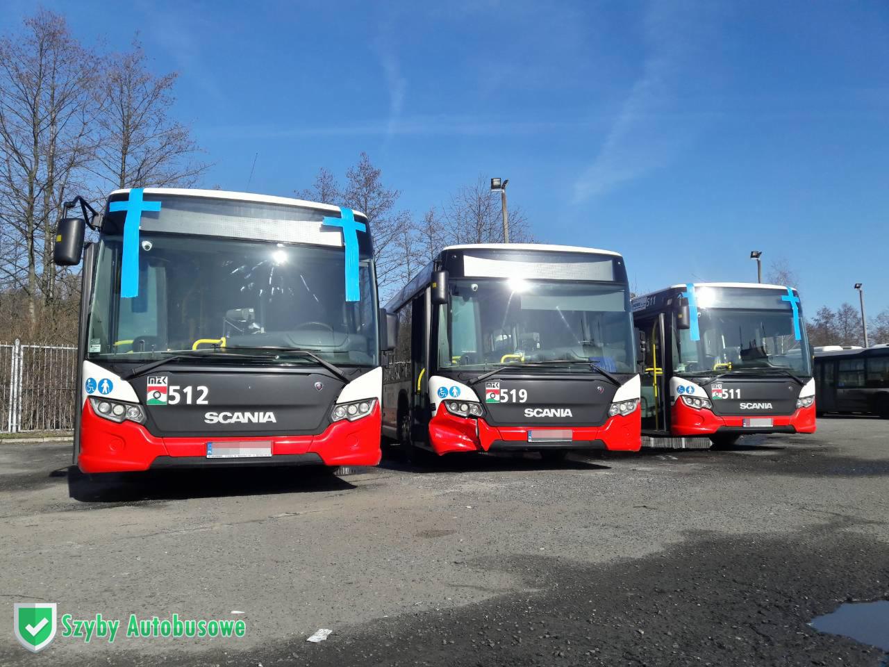 Trzy nowe autobusy Scania w kolorach miejskich, zaparkowane na placu, z niebieską taśmą zabezpieczającą na przedniej szybie jednego z nich.