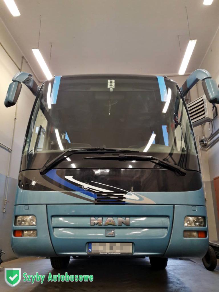 Przód niebieskiego autobusu marki MAN w warsztacie, z taśmą malarską na szybie, sugerującą wymianę lub naprawę szyby przedniej.