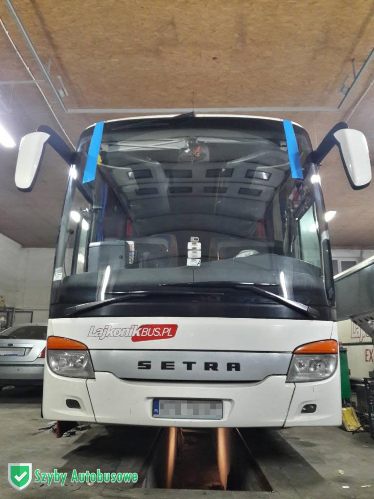 Przód białego autobusu Setra na stanowisku warsztatowym z otwartym kanałem, z taśmą malarską zabezpieczającą szybę. Widoczne logo LajkonikBUS.PL.
