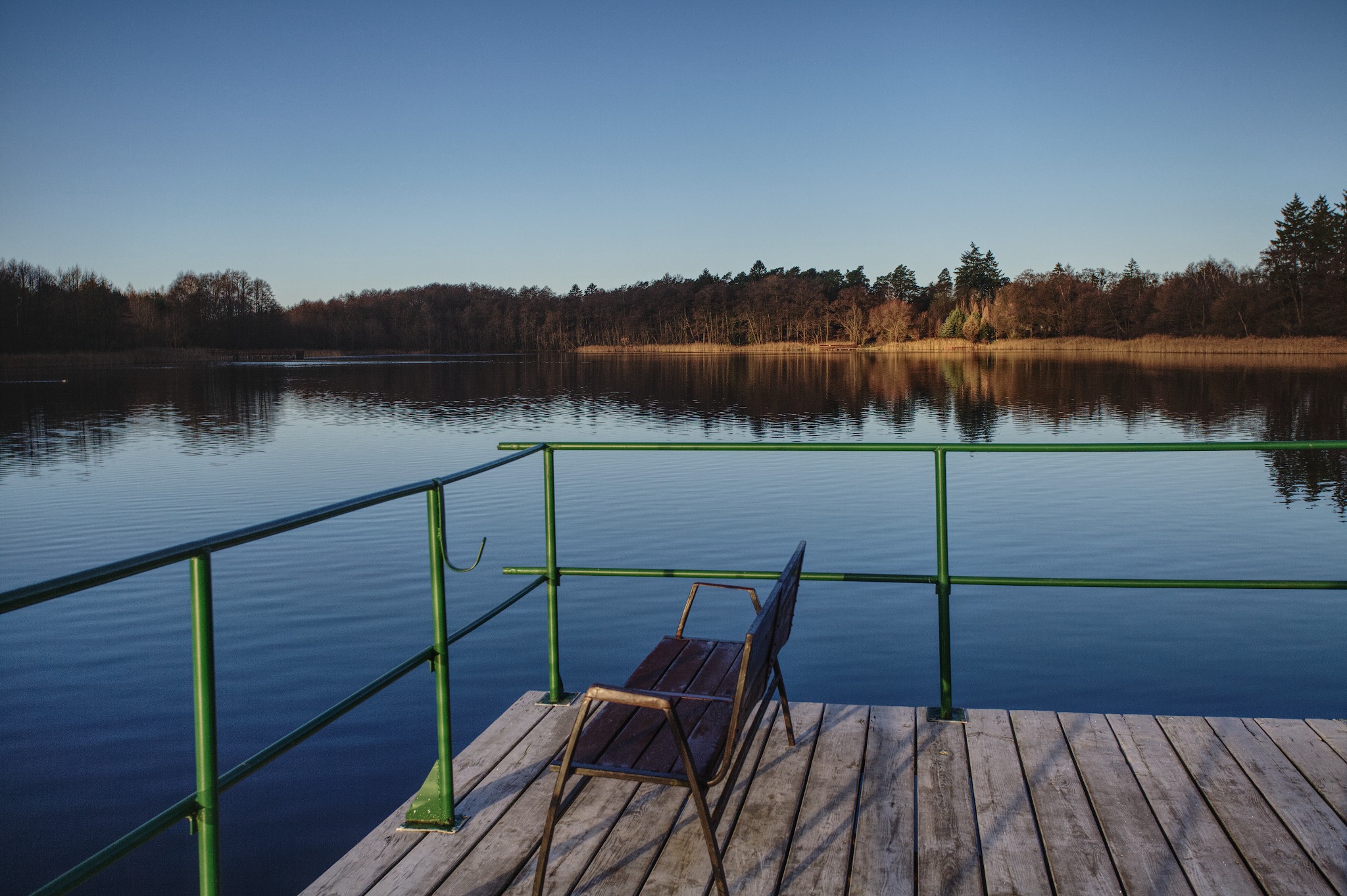 Drewniany pomost z zieloną barierką i ławką, widok na spokojne jezioro i las w oddali.