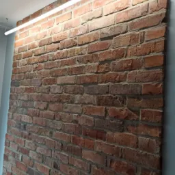 Ściana z czerwonej cegły z widocznymi spoinami i zamontowanym poziomym oświetleniem LED, surowy styl wykończenia wnętrza.