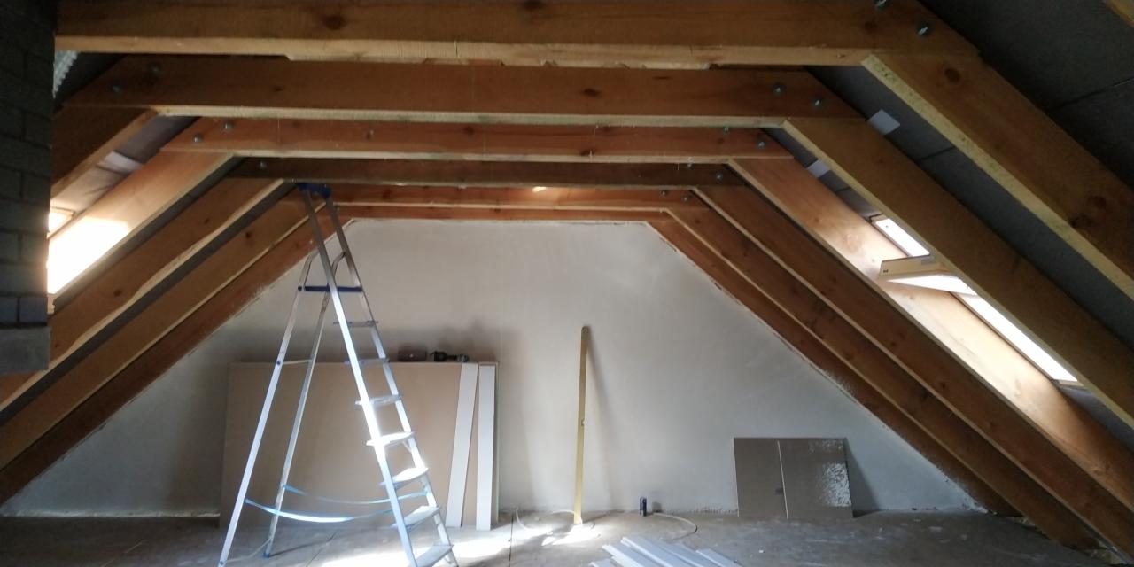 Poddasze w trakcie adaptacji: widoczne drewniane krokwie, metalowa drabina oparta o ścianę, płyty gipsowo-kartonowe i listwy przypodłogowe przygotowane do montażu, okno dachowe w skosie.