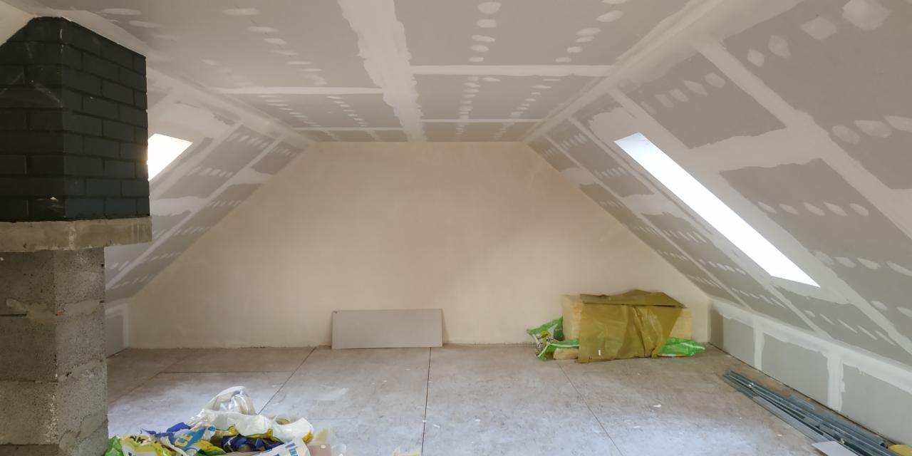 Poddasze w trakcie prac wykończeniowych: ściany i sufit pokryte płytami gipsowo-kartonowymi, widoczne łączenia wymagające szpachlowania, ceglany komin pomalowany na czarno, okno dachowe w skosie...