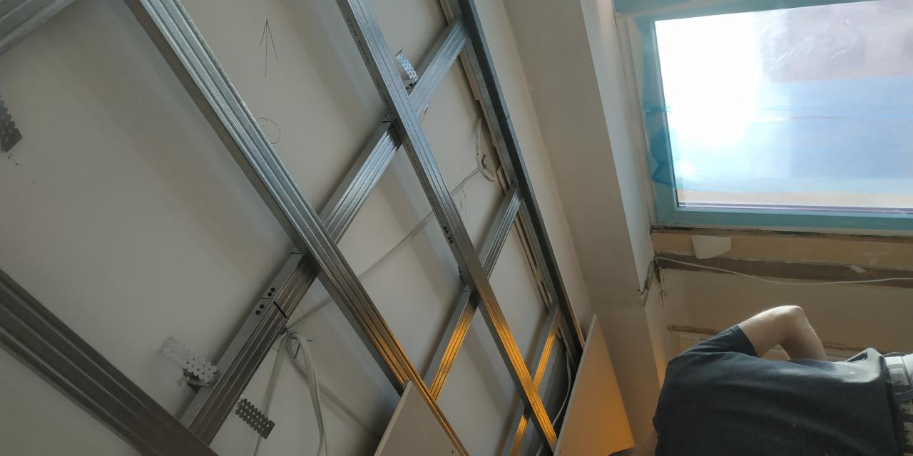 Montaż metalowego rusztu pod płyty gipsowo-kartonowe na poddaszu, widoczne profile, uchwyty montażowe i częściowo zamontowana płyta, okno dachowe w tle.