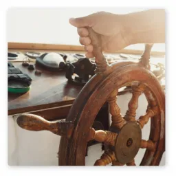 Dłoń na drewnianym kole sterowym jachtu w słoneczny dzień, widoczne instrumenty nawigacyjne w tle.