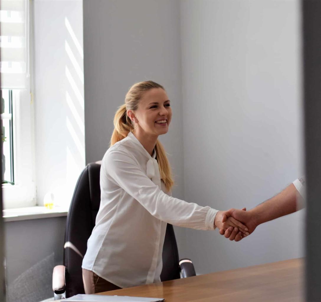 Uśmiechnięta blondynka w białej bluzce z kokardą wita się z klientem przy drewnianym biurku w jasnym biurze, widok częściowo zasłonięty przez framugę drzwi.
