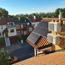 Widok z góry na dach domu z panelami słonecznymi i dwoma klimatyzatorami marki AUX zamontowanymi na ceglanej ścianie komina, w tle zabudowa miejska.