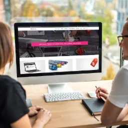 Dwie osoby przeglądają stronę internetową agencji reklamowej 'Doneta' na monitorze komputera; widoczne elementy strony to oferta reklamy, stron internetowych, druku i poligrafii.