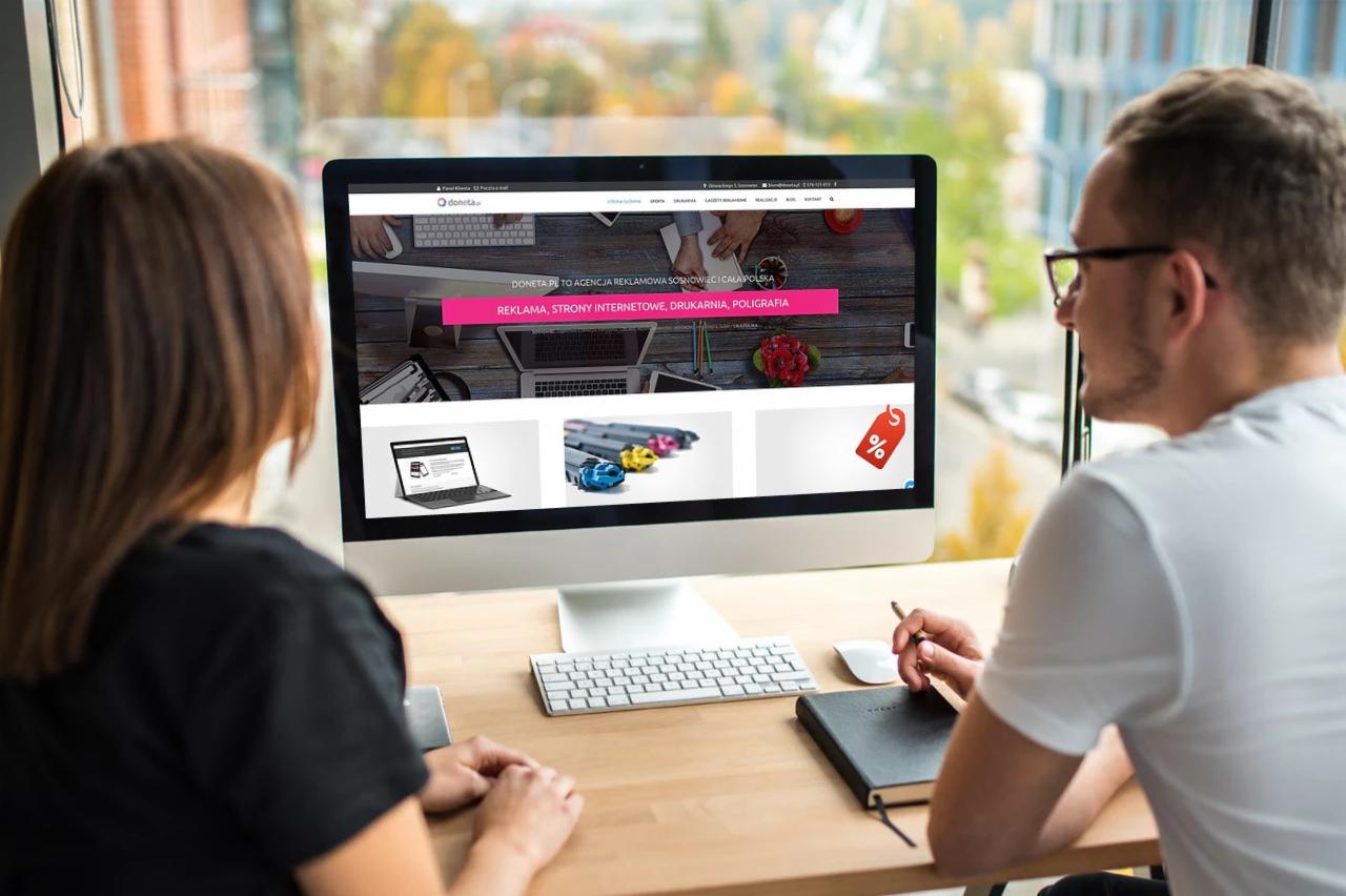 Dwie osoby przeglądają stronę internetową agencji reklamowej 'Doneta' na monitorze komputera; widoczne elementy strony to oferta reklamy, stron internetowych, druku i poligrafii.