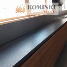 Długi blat kamienny w ciemnym odcieniu szarości, zamontowany w kuchni pod oknem, z widocznym fragmentem okna i logo firmy Kominki Vento nałożonym na zdjęcie.