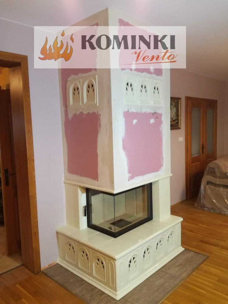 Biały kominek kaflowy w trakcie montażu, z widocznymi śladami gipsu na ścianie, ozdobne kafle z motywem gotyckim, przeszklone drzwiczki paleniska.