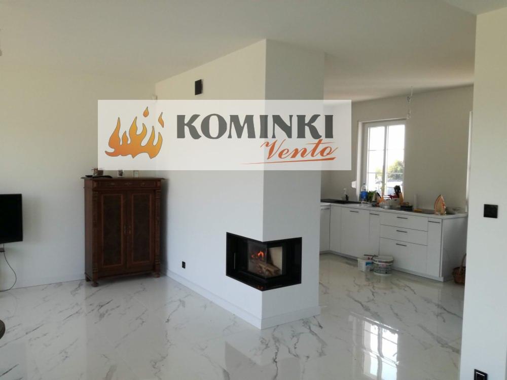 Nowoczesny kominek narożny z paleniskiem w jasnym salonie z marmurową podłogą, widoczna część kuchni w tle.