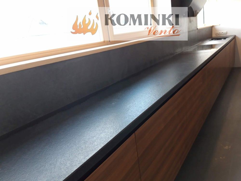 Długi blat kamienny w ciemnym odcieniu szarości, zamontowany w kuchni pod oknem, z widocznym fragmentem okna i logo firmy Kominki Vento nałożonym na zdjęcie.