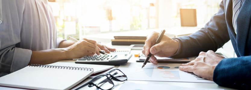 Dwóch analityków finansowych analizuje wykresy i obliczenia przy użyciu kalkulatora, długopisu i notatnika w jasnym biurze.