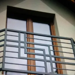 Szara, metalowa balustrada balkonowa z geometrycznym wzorem na tle okna w brązowej ramie i jasnej elewacji budynku.