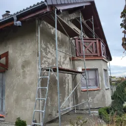 Dom jednorodzinny w trakcie tynkowania maszynowego, widoczne rusztowanie, surowy tynk na elewacji, zabezpieczone okna folią malarską, fragment ogrodu z iglakami.