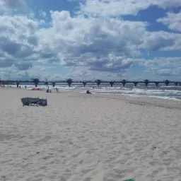 Szeroki kadr plaży z widokiem na molo z platformami widokowymi, osoby w oddali, parawan plażowy po lewej stronie, błękitne niebo z chmurami.