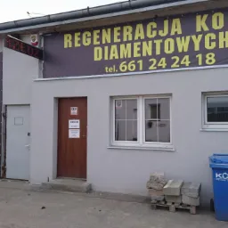 Fasada budynku firmy z banerami reklamowymi oferującymi regenerację koron diamentowych, wynajem narzędzi ogrodniczych, serwis i sprzedaż, z widocznymi niebieskimi pojemnikami na odpady komunalne.